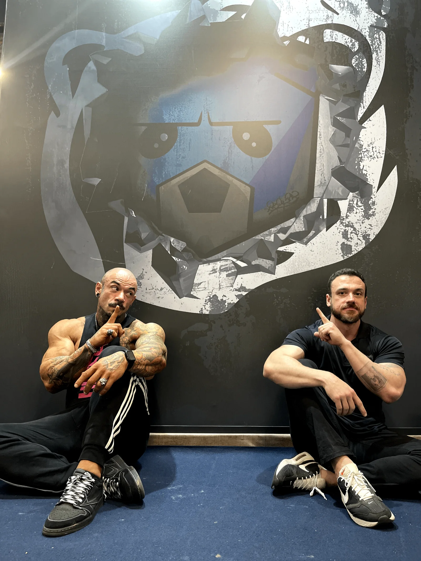 Will Jardim e atleta parceiro sentados diante de mural gigante do urso da Under Labz
