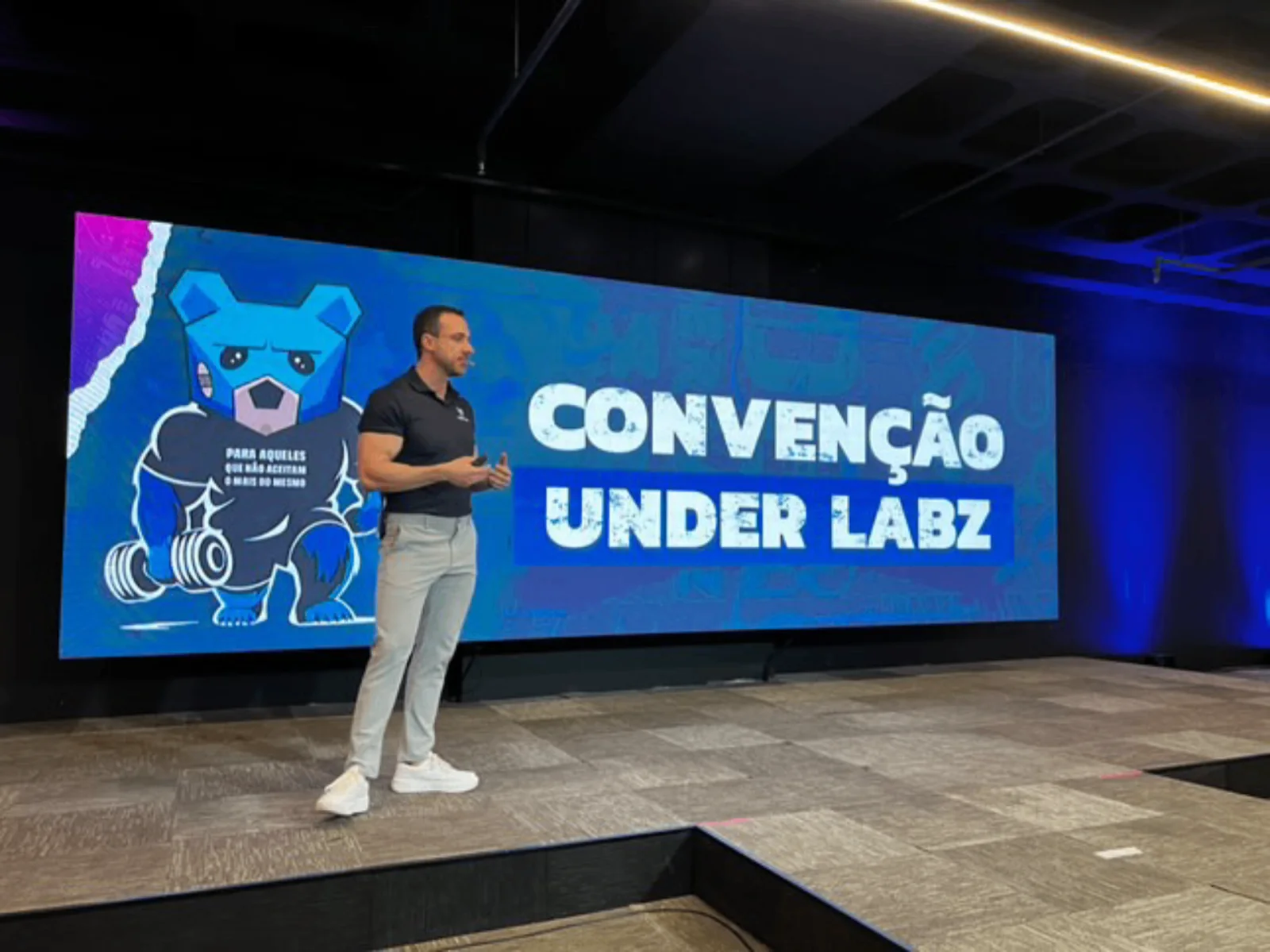 Will Jardim palestrando no palco da Convenção Under Labz diante de painel de LED com o mascote urso da marca
