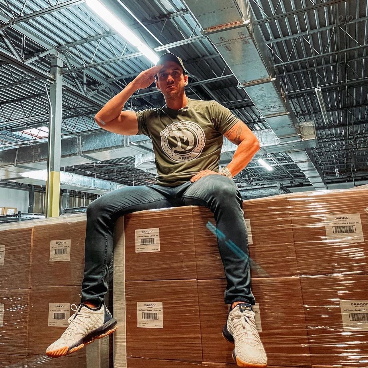 Will Jardim com camiseta Dragon Army sentado sobre caixas da Dragon Pharma em um galpão